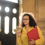 A young woman in a library carrying a red binder, bathed in sunlight.